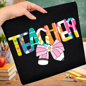 Pu&ograve; includere: T-shirt nera con la scritta "TEACHER" in lettere colorate e decorate. Un fiocco rosa con un fiore sorridente e dettagli di matita &egrave; sotto la scritta. La maglietta &egrave; tenuta in mano.