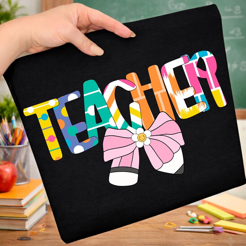 Pu&ograve; includere: T-shirt nera con la scritta "TEACHER" in lettere colorate e decorate. Un fiocco rosa con un fiore sorridente e dettagli di matita &egrave; sotto la scritta. La maglietta &egrave; tenuta in mano.