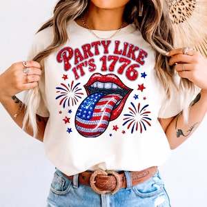 May include: Cream-colored t-shirt with a graphic that says "PARTY LIKE IT'S 1776." The graphic features red lips, a tongue with a US flag design, red and blue stars, and fireworks.