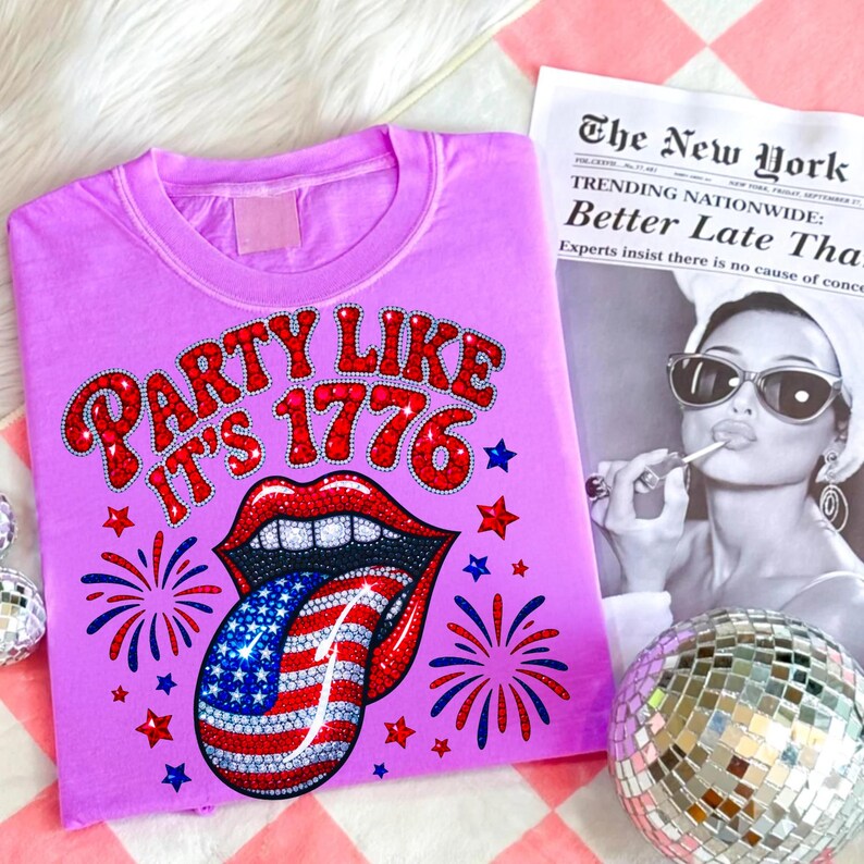 May include: A lilac-colored t-shirt with the phrase "PARTY LIKE IT'S 1776" in red and white lettering, featuring a graphic of a mouth with a tongue designed as the American flag. Fireworks and stars surround the graphic.