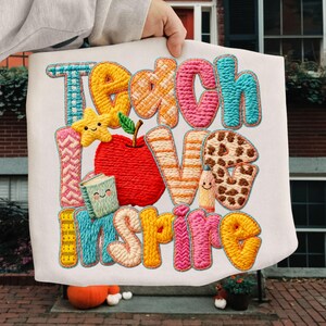 May include: A white fabric item featuring the embroidered words "Teach Love Inspire" in vibrant, textured lettering. The design incorporates a red apple, a smiling star, a book, and a pencil, creating a playful, educational theme.