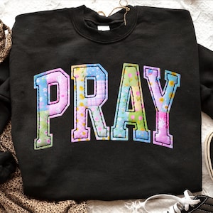 Może przedstawiać: Czarna bluza z napisem "PRAY" w kolorowym patchworku. Litery są obrysowane na biało i wypełnione kwiatowymi i wzorzystymi tkaninami w odcieniach różu, błękitu, zieleni i żółci. Bluza ma okrągły dekolt.