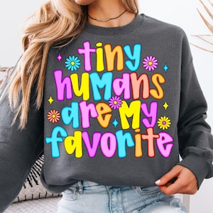以下が含まれることがあります： 「tiny humans are my favorite」の文字がカラフルなバブルレターでプリントされた、濃いグレーのスウェットシャツ。デザインには花と星が含まれています。スウェットシャツはカジュアルなスタイルです。