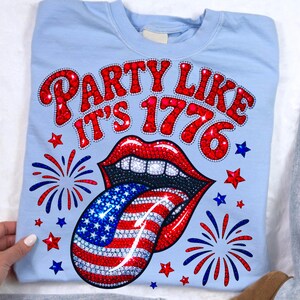 May include: Light blue sweatshirt with the text "PARTY LIKE IT'S 1776" in red, with a red-lipped mouth and a tongue designed like the American flag. Red, white, and blue stars and fireworks complete the design.