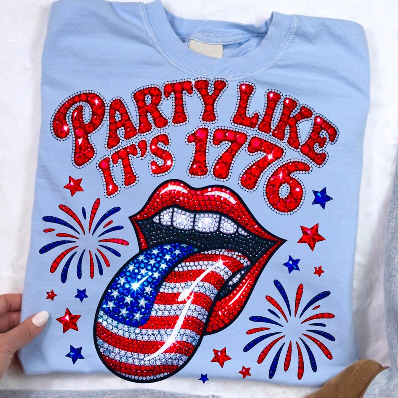 May include: Light blue sweatshirt with the text "PARTY LIKE IT'S 1776" in red, with a red-lipped mouth and a tongue designed like the American flag. Red, white, and blue stars and fireworks complete the design.