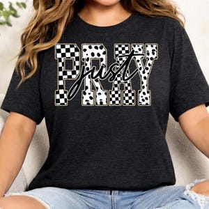 Könnte beinhalten: Dunkelgraues T-Shirt mit den Wörtern "PRAY" und "just". Das Wort "PRAY" ist in einem schwarz-weiß karierten und Dalmatiner-Muster gehalten. Das Wort "just" ist in Schreibschrift. Kurzärmlig.