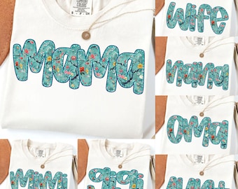 May include: Cream-colored t-shirts with floral text designs. The shirts feature the words "Mama", "Wife", "Nana", "Oma", "Mimi", "Gigi", and "Aunt" in a teal, floral font. Each shirt has a gold necklace.
