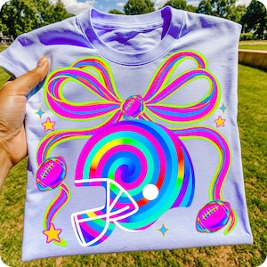 May include: A light purple t-shirt featuring a colorful graphic design. The design includes a football helmet, footballs, a large bow, stars, and a spiral design in shades of blue, green, pink, and yellow.
