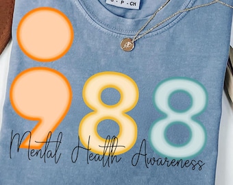 May include: A light blue t-shirt with the words "Mental Health Awareness" and the numbers "988" in orange, yellow, and teal. A gold necklace with a small pendant is also visible.