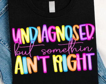 Pode incluir: Camiseta preta com a frase "UNDIAGNOSED but somethin AIN'T RIGHT" em letras coloridas. As letras são rosa, amarelas, azuis e laranjas. A camiseta está dobrada sobre uma superfície de ganga.