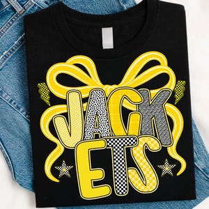 May include: Black t-shirt with the word "JACKETS" in a yellow bow design. The letters are in various patterns, including polka dots, plaid, and checkerboard. Two stars and lightning bolts accent the design.