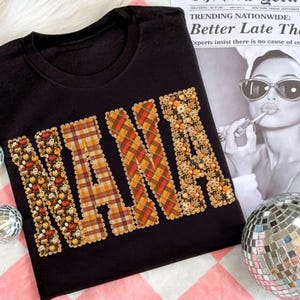 May include: Black t-shirt featuring the word "NANA" in large, decorative letters. The letters are filled with autumn-themed patterns, including pumpkins, flowers, and plaid. The shirt is displayed on a pink and white checkered surface.