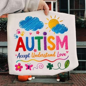 Puede incluir: Tela blanca con bordados coloridos. La palabra "AUTISMO" está bordada en rojo, azul, verde, morado y naranja. Debajo, la frase "Accept Understand Love" está escrita en cursiva. El diseño incluye nubes, un sol, flores y una mariposa.