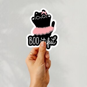 May include: A black cat sticker with a pink bow and tutu. The sticker has the words "BOO-tiful" in a playful font. The cat has white eyes and whiskers. The sticker is held by a hand.