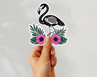 May include: A die-cut sticker featuring a black flamingo silhouette with a visible white ribcage skeleton design. The flamingo is positioned above two vibrant pink hibiscus flowers and several dark green tropical palm leaves.