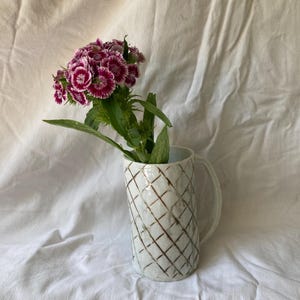 Può includere: Un vaso di ceramica bianca con un motivo a griglia marrone contiene un bouquet di fiori rosa e bianchi con foglie verdi. Il vaso ha un manico ed è posto su uno sfondo di tessuto bianco.