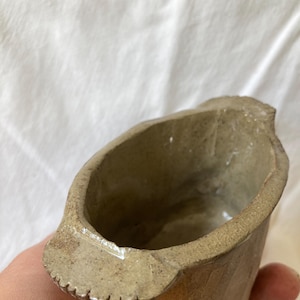 May include: A handmade, small, ceramic bowl with a unique, irregular shape. The bowl is a muted, earthy brown color with a textured surface and a small spout. The rim has a decorative, notched edge. The interior is a lighter, glazed color.