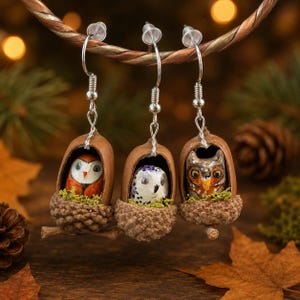 May include: Three pairs of handcrafted owl earrings. Each earring features a miniature owl figurine nestled inside a brown acorn shell. The owls have colorful details, and the earrings hang from silver-colored hooks.