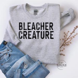 May include: A light gray sweatshirt with the words "BLEACHER CREATURE" printed in black. The sweatshirt is folded with a pair of blue jeans. A sprig of decorative gold-colored berries is on the right.