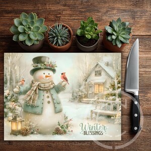 May include: A decorative cutting board with a winter scene featuring a snowman, cardinals, and a snow-covered house. The board has the text "Winter Blessings". A stainless steel knife and potted succulents are also in the image.