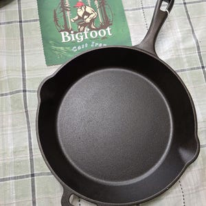 Puede incluir: Una sart&eacute;n de hierro fundido negro con un mango largo y un asa auxiliar. Un pa&ntilde;o verde con el logotipo de Bigfoot Cast Iron est&aacute; en el fondo. La sart&eacute;n est&aacute; sobre una superficie a cuadros verdes y blancos.