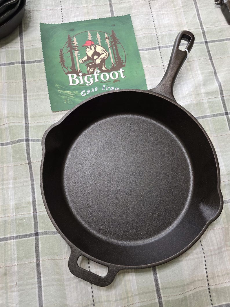 Puede incluir: Una sart&eacute;n de hierro fundido negro con un mango largo y un asa auxiliar. Un pa&ntilde;o verde con el logotipo de Bigfoot Cast Iron est&aacute; en el fondo. La sart&eacute;n est&aacute; sobre una superficie a cuadros verdes y blancos.