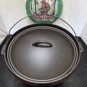 May include: A black cast iron Dutch oven with a lid and handle. The lid has a small handle. The pot has a handle for carrying. The background has a green circle with the words "Bigfoot Cast Iron LLC" and an illustration of Bigfoot.