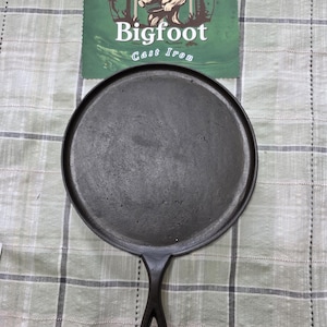 May include: A black cast iron griddle with a handle. The griddle is round with a flat cooking surface. The handle has a hole for hanging. A green sign with the text "Bigfoot Cast Iron" and an illustration of Bigfoot is in the background.