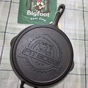 Puede incluir: Sart&eacute;n de hierro fundido negro con el texto en relieve "PIT BOSS" y un dise&ntilde;o de chef de dibujos animados. La sart&eacute;n tiene un asa y un peque&ntilde;o lazo en el lado opuesto. Una etiqueta verde con el texto "Bigfoot Cast Iron" es visible.