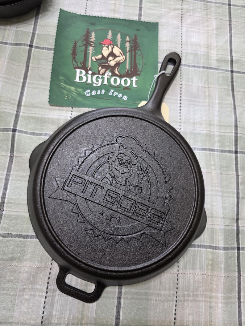 Puede incluir: Sart&eacute;n de hierro fundido negro con el texto en relieve "PIT BOSS" y un dise&ntilde;o de chef de dibujos animados. La sart&eacute;n tiene un asa y un peque&ntilde;o lazo en el lado opuesto. Una etiqueta verde con el texto "Bigfoot Cast Iron" es visible.