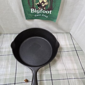 May include: A black cast iron skillet with a long handle. Above the skillet is a green banner with the words "Bigfoot Cast Iron" and an illustration of Bigfoot cooking.
