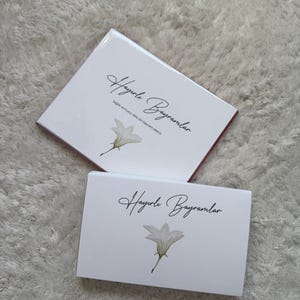 Può includere: Due scatole regalo bianche con il testo "Hayırlı Bayramlar" in corsivo nero, ognuna con un'illustrazione di fiore bianco. Le scatole sono rettangolari e poggiano su una superficie bianca strutturata.