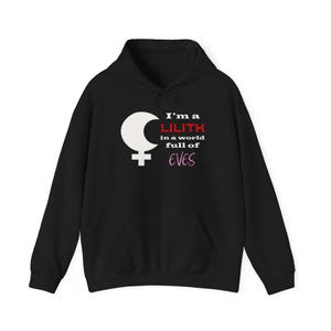 May include: Black hooded sweatshirt with a white crescent moon and cross symbol. The text reads "I'm a LILITH in a world full of EVES" in white, red, and pink lettering. The sweatshirt has a front pocket and a drawstring hood.