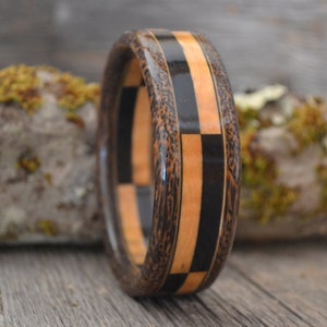 May include: A wooden ring featuring a black and brown chequered pattern. The ring is crafted from various types of wood, including a light wood with a grain pattern.