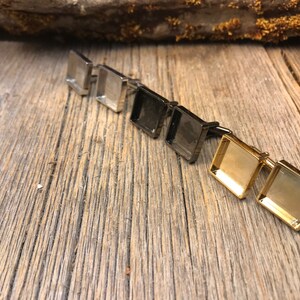 Puede incluir: Cuatro pares de gemelos con inserciones de vidrio cuadradas. Los gemelos son de plata, negro y oro.