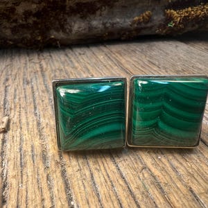 May include: A pair of square cufflinks featuring polished malachite stones with a vibrant green color and unique striped patterns. The stones are set in a silver-colored metal frame, showcasing a classic design.