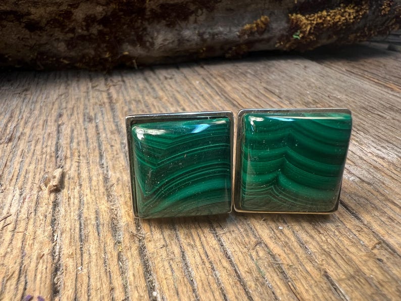 May include: A pair of square cufflinks featuring polished malachite stones with a vibrant green color and unique striped patterns. The stones are set in a silver-colored metal frame, showcasing a classic design.