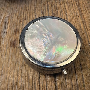 Könnte beinhalten: Ein rundes, silberfarbenes Metalldöschen mit einer kreisförmigen Einlage aus schillerndem Perlmutt auf dem Deckel. Das Kompaktgehäuse hat eine glatte, polierte Oberfläche und einen kleinen, hervorstehenden Metallverschluss an der Seite.