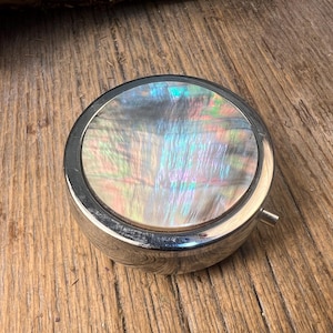 May include: A circular, silver-coloured pill box with a mother-of-pearl lid. The iridescent surface of the lid reflects blue, green, and pink hues. The pill box is on a wooden surface.