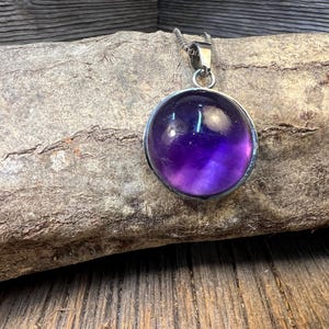 Könnte beinhalten: Eine runde, lila Amethyst-Anhänger-Halskette mit einer silberfarbenen Kette. Der Edelstein ist in einer silberfarbenen Lünette gefasst. Der Anhänger wird auf einem Stück verwittertem Holz präsentiert.
