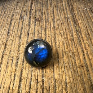 May include: A circular, silver ring with a polished, blue gemstone exhibiting iridescent colours. The stone has a vibrant play of colours, set against a rustic, wooden background. The ring is a piece of jewellery.