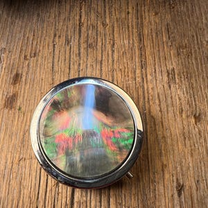 May include: A circular, silver compact mirror with an iridescent, shimmering surface. The surface has a mix of colours, including green, red, and blue. The compact is set against a rustic, wooden background.