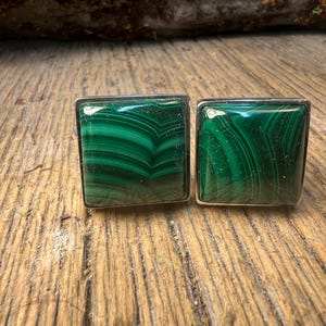May include: A pair of square cufflinks featuring polished malachite stones with a vibrant green color and unique swirling patterns. The cufflinks are set in a silver-colored metal frame, showcasing the natural beauty of the gemstone.