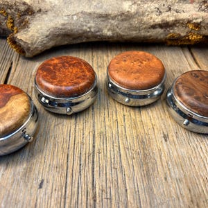 May include: Four round pill cases with polished silver-tone metal bases and lids featuring inlaid natural wood burl with distinct swirling grain patterns in shades of amber, brown, and tan, arranged in a semi-circle on a weathered wooden surface.