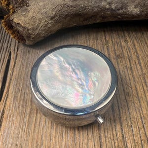 May include: A round, silver-toned metal pill box or compact featuring a circular inlay of iridescent mother-of-pearl on the lid. The surface of the inlay displays swirling patterns of white, pink, and blue hues with a polished, reflective finish.
