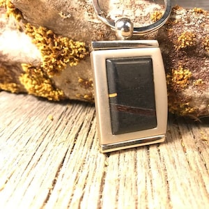 May include: A silver key chain with a black rectangular charm featuring a wood inlay. The charm has a thin gold line running through it.