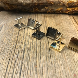 May include: A collection of square cufflinks in silver, black, and gold tones. The cufflinks have a rectangular post and a square face with a geometric design. They are arranged on a weathered wooden surface.