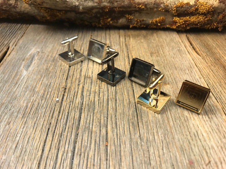 May include: A collection of square cufflinks in silver, black, and gold tones. The cufflinks have a rectangular post and a square face with a geometric design. They are arranged on a weathered wooden surface.