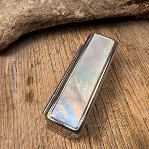 May include: A rectangular metal lipstick case featuring a polished silver-tone finish and an inlaid panel of iridescent mother-of-pearl. The surface of the inlay displays a swirling pattern of soft pink, blue, and white hues with a glossy finish.