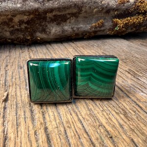 May include: Two square cufflinks with a silver border. The cufflinks are made of polished malachite, displaying a vibrant green color with swirling patterns. The cufflinks are displayed on a wooden surface.